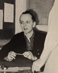 Black and white photo of Ron Underwood teaching in the 1960s, seated at a desk and engaging with a student during his early years as a speech and debate coach at Downey High School.